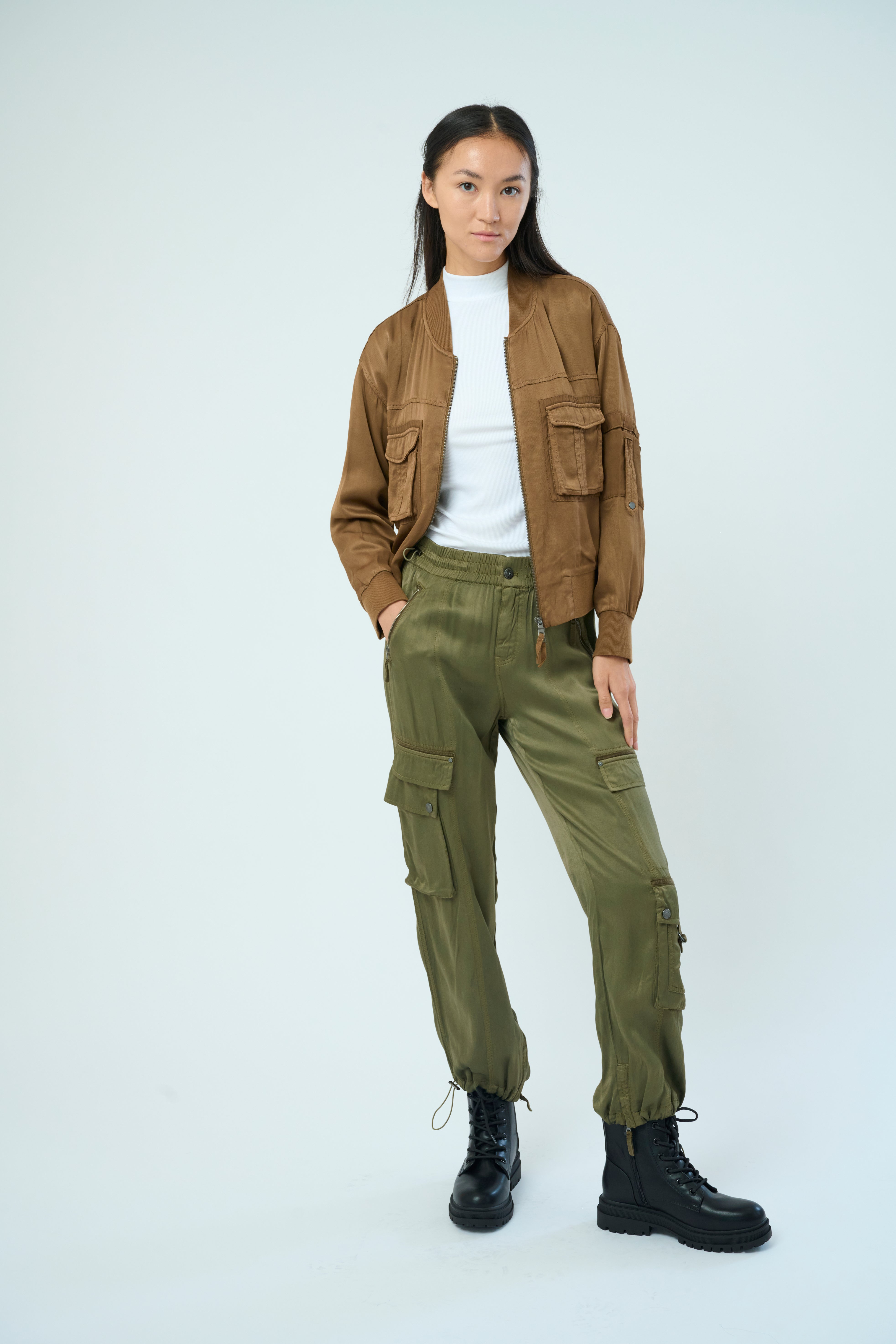 A tall female model with long black hair confidently poses with hands in pockets, wearing our cheetah colored Dazie flight jacket with a brown zipper from Marrakech Clothing.