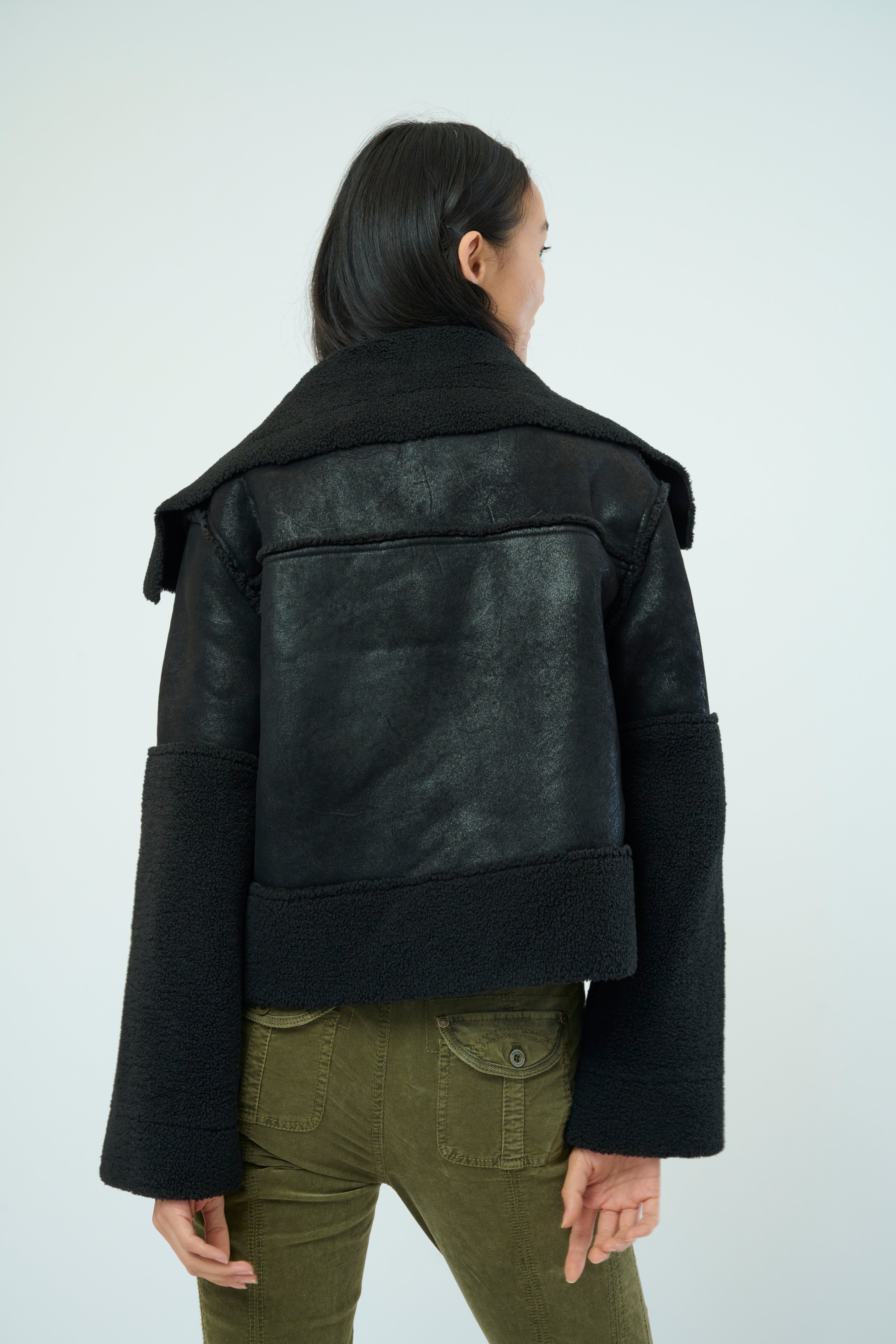 Back view of a tall female model with long black hair confidently posing wearing our black colored Trisha Bonded Distressed Suede Sherpa Jacket from Marrakech Clothing.