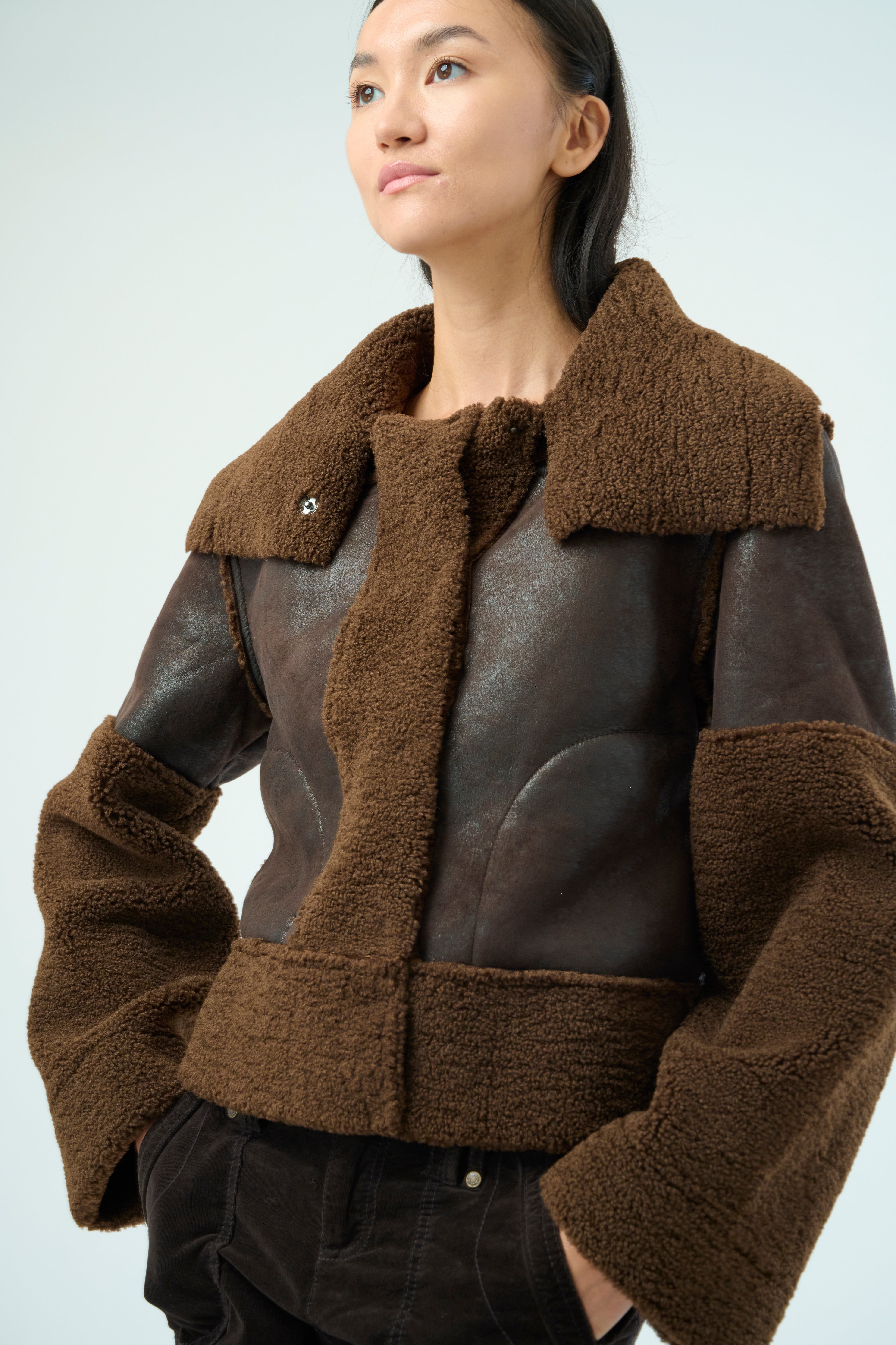 Close up view of a tall female model with long black hair confidently posing wearing our dark chocolate colored Trisha Bonded Distressed Suede Sherpa Jacket from Marrakech Clothing.